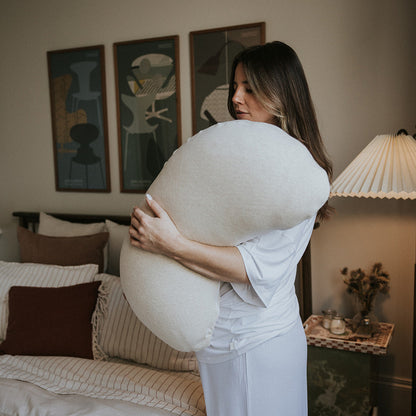 A woman in a white lounge set stands in a bedroom, hugging a large, rounded off-white lumbar pillow. The bedroom features neutral beige and brown tones, with abstract art prints on the wall, a bedside table with a lamp, and a bed with striped bedding.
