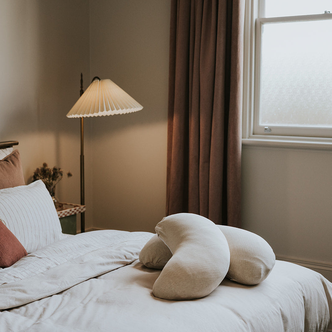 A cozy bedroom scene with a neatly made bed in soft white bedding, styled with striped and rust-colored pillows. Two cream crescent-shaped lumbar pillows rest on the bed, next to a lit floor lamp and a window with brown curtains.